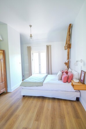Een slaapkamer met een houten vloer, een bed met wit bedlinnen en roze kussens, een houten nachtkastje, een wandlamp en een raam met gordijnen.