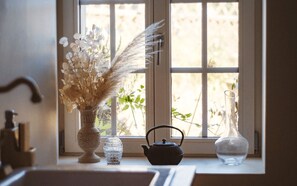 Une fenêtre donnant sur un paysage verdoyant, un vase contenant des fleurs séchées, un vase en verre transparent et une théière noire posée sur le rebord de la fenêtre.