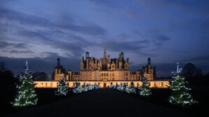 Un edificio majestuoso e histórico con múltiples torres y una fachada simétrica, rodeado de árboles de Navidad iluminados.