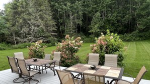 An outdoor deck with a table and chairs, surrounded by lush greenery and flowering bushes.