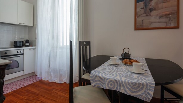 Une cuisine de taille réduite avec des meubles blancs, une machine à café et une table à manger recouverte d’une nappe à motifs.
