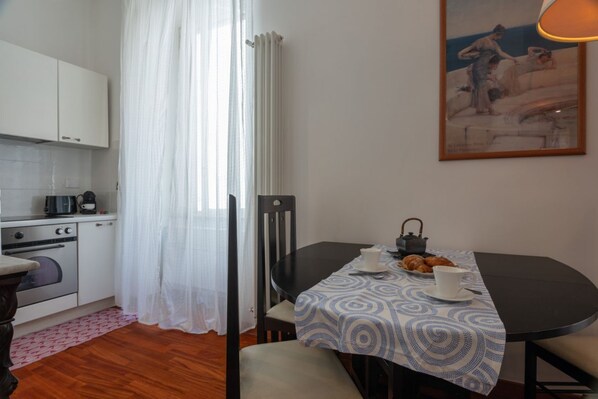 Une cuisine de taille réduite avec des meubles blancs, une machine à café et une table à manger recouverte d’une nappe à motifs.