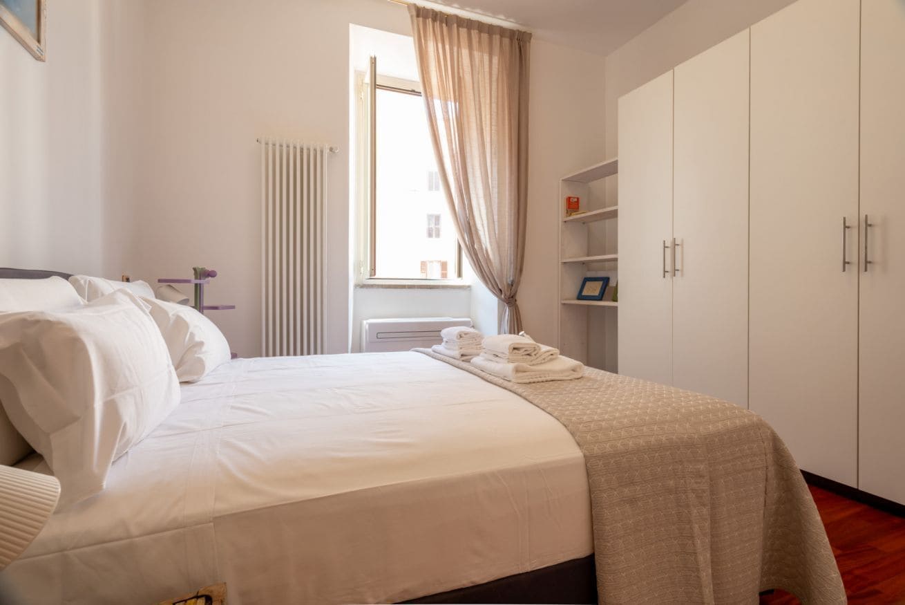 Un letto rifatto con lenzuola bianche, una finestra con tende, un termosifone e un armadio con maniglie.