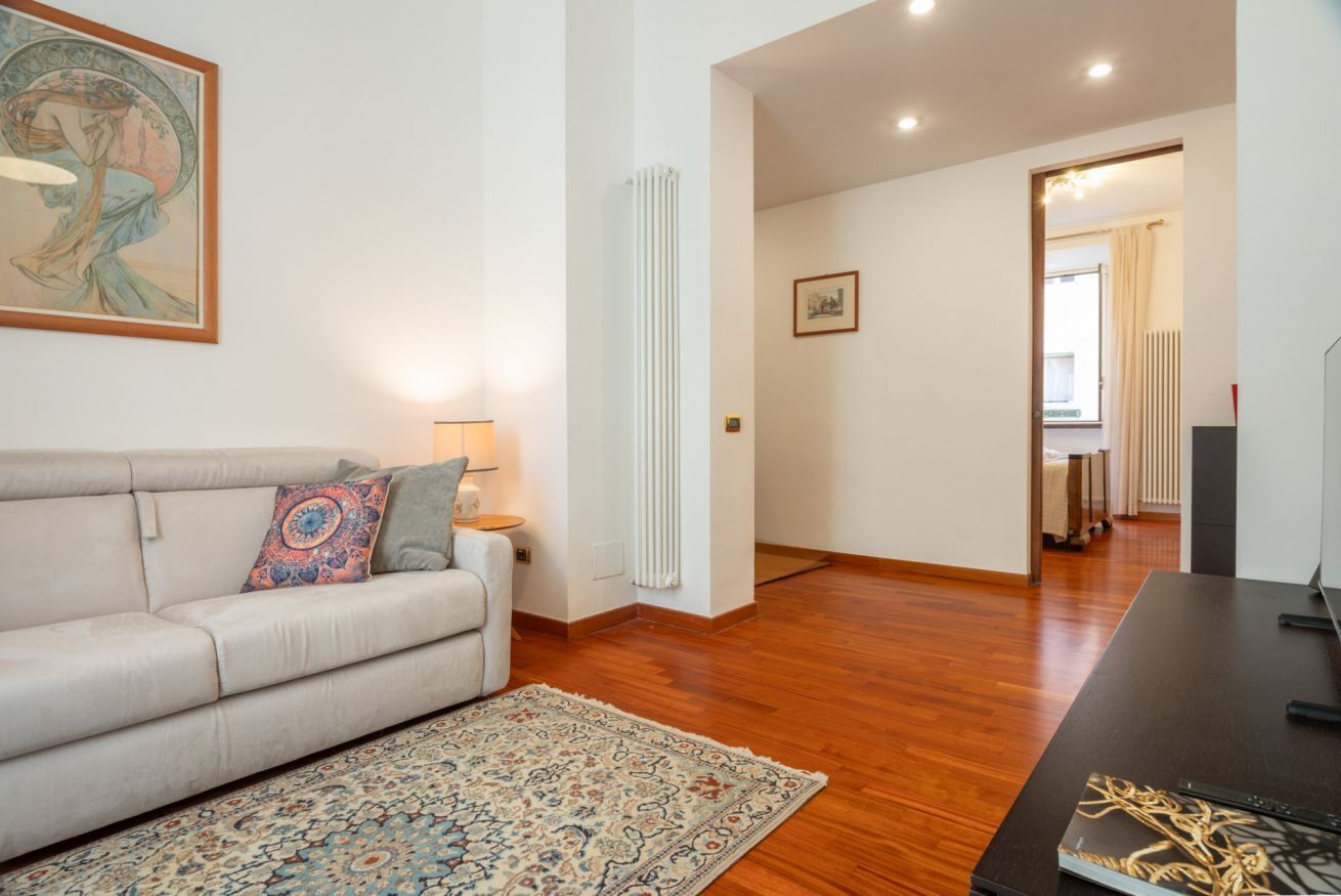 A living room with a sofa, a patterned rug, a painting, a radiator, and a doorway leading to another room.