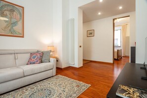 Un salón con sofá, alfombra estampada, un cuadro, radiador y un pasillo que conduce a otra habitación.