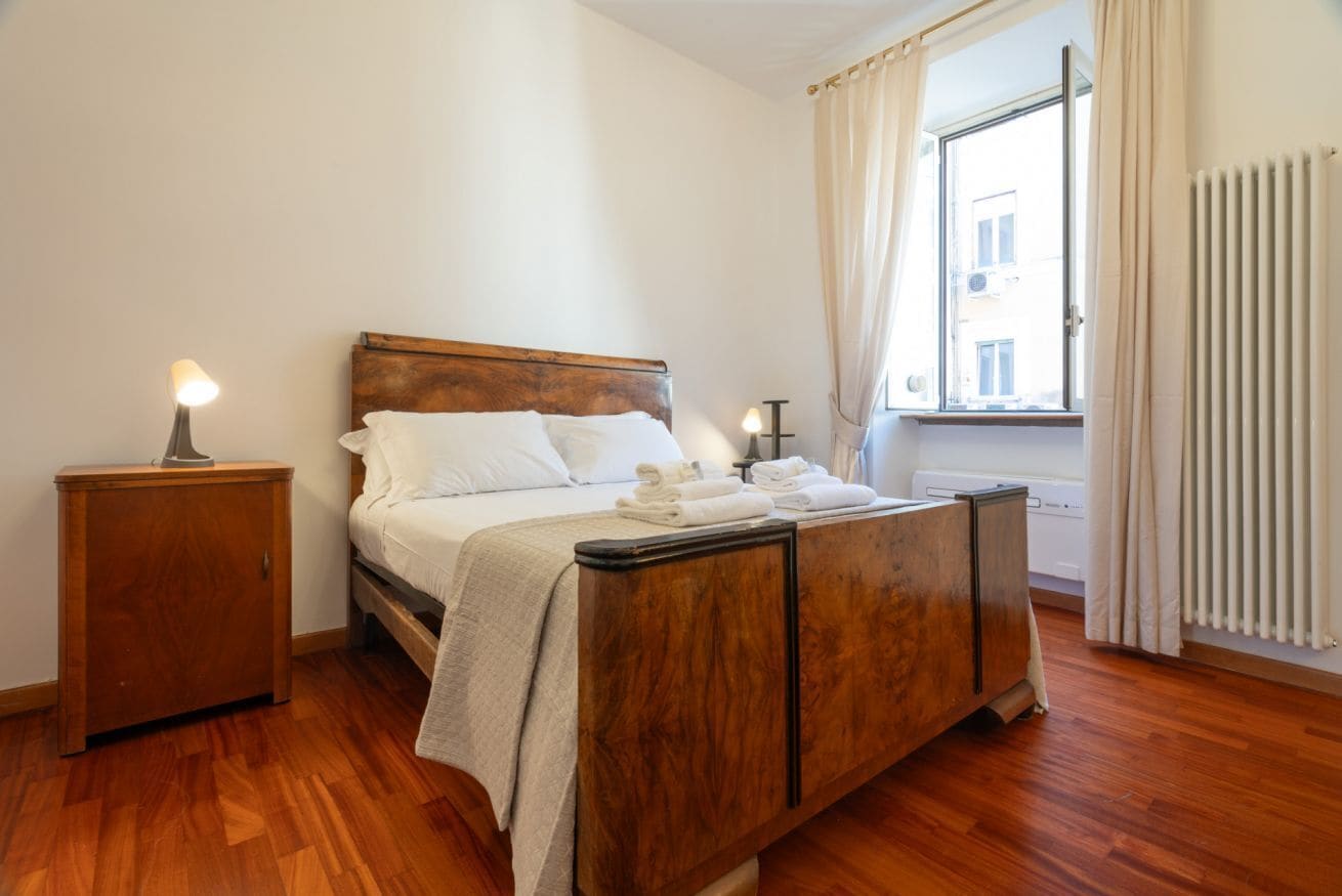 Une chambre à coucher avec un lit en bois, une table de chevet, une fenêtre avec des rideaux et un radiateur.