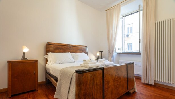A bedroom with a wooden bed, a nightstand, a window with curtains, and a radiator. - Casa Novecento Prati (Roma)