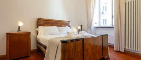 A bedroom with a wooden bed, a nightstand, a window with curtains, and a radiator.