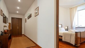 A hotel room with a wooden floor, a bed with a white bedspread, a bedside table with a lamp, and a window with curtains. - Casa Novecento Prati (Roma)