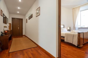 A hotel room with a wooden floor, a bed with a white bedspread, a bedside table with a lamp, and a window with curtains.