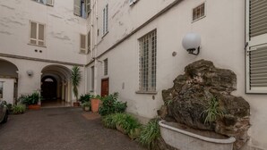 A courtyard with a stone fountain, potted plants, and a building with a tiled roof and arched entrance. - Casa Novecento Prati (Roma)