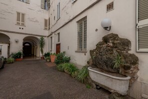 A courtyard with a stone fountain, potted plants, and a building with a tiled roof and arched entrance.