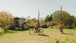 Dos personas montando en bicicleta por un área con césped, con un edificio pequeño y árboles al fondo.
