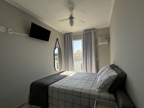 Een slaapkamer met een bed, een plafondventilator, een tv aan de muur en een raam met gordijnen.