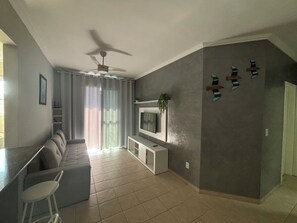 Een moderne woonkamer met een grijze bank, een tv die aan een witte console is bevestigd, een plafondventilator en een groot raam met gordijnen.