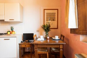 Une petite cuisine avec une table à manger, des chaises, un ordinateur et un panier de fruits.
