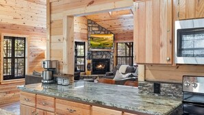 A log cabin interior with a stone fireplace, wooden cabinets, and a kitchen area.