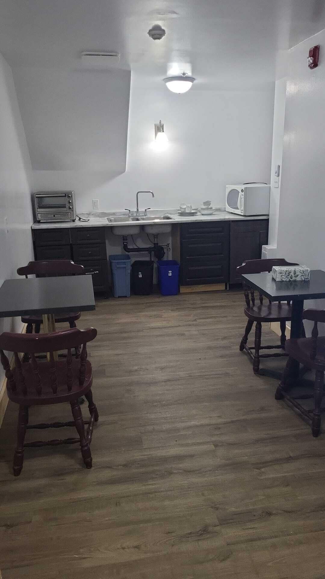 Una cocina compacta con un lavamanos, un microondas y un tostador. En primer plano, hay dos sillas de madera y una mesa.