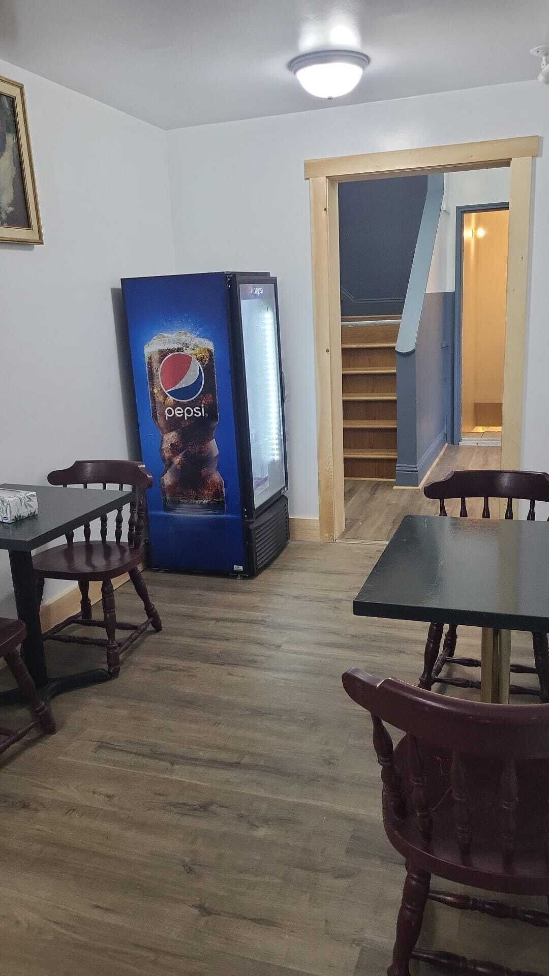 Et værelse med trægulv, to træborde og stole. Et Pepsi køleskab er synligt.