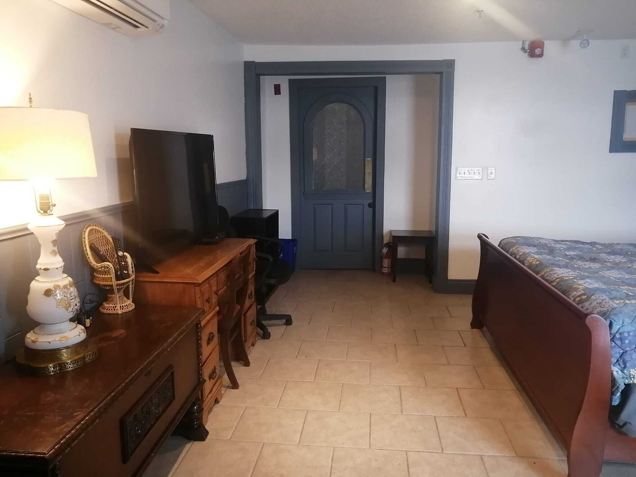 Une chambre à coucher avec une commode en bois, un téléviseur, un lit et une porte donnant sur une autre pièce.