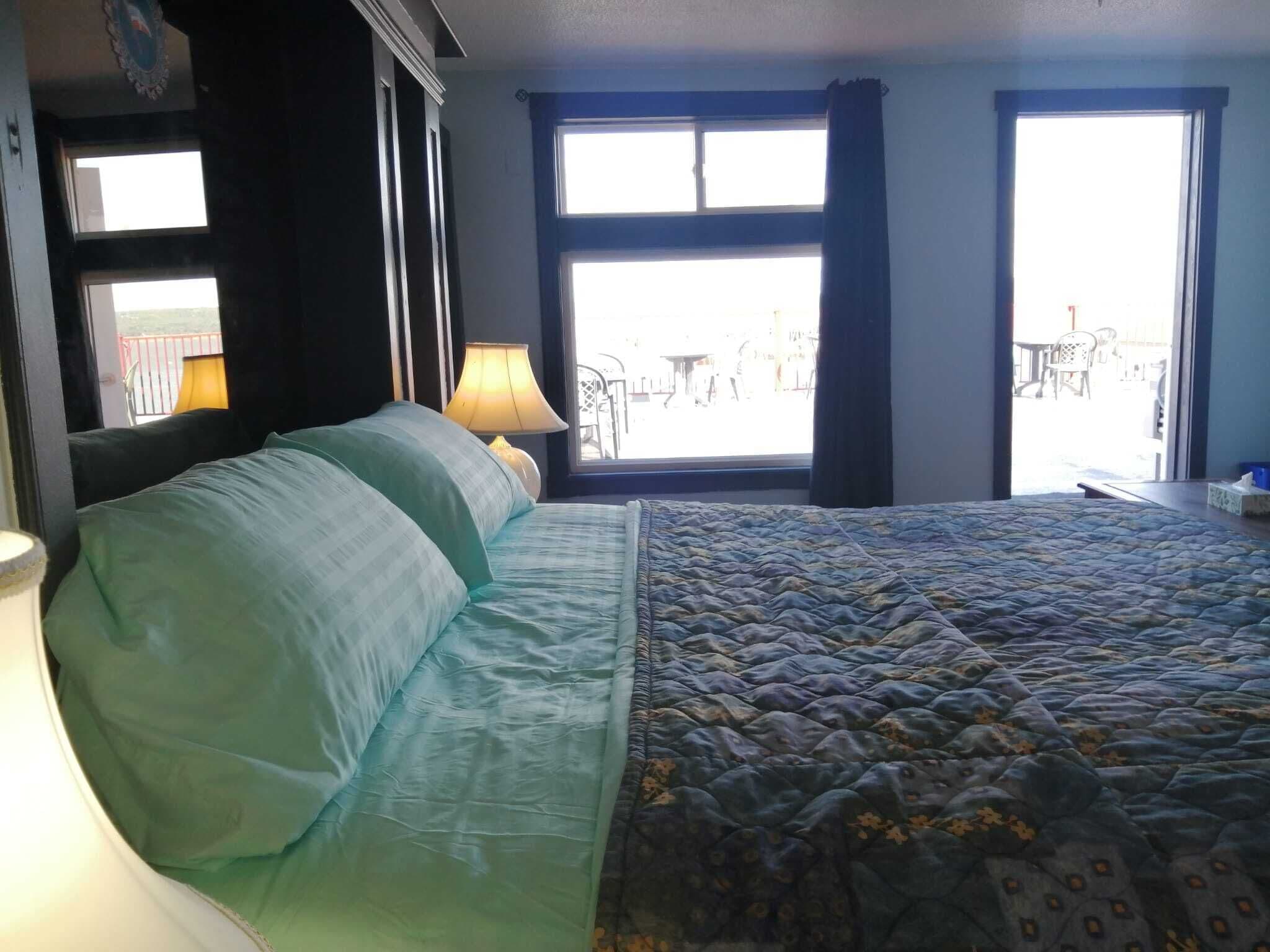Una habitación de hotel con una cama, una mesita de noche con lámpara y vistas al balcón exterior.