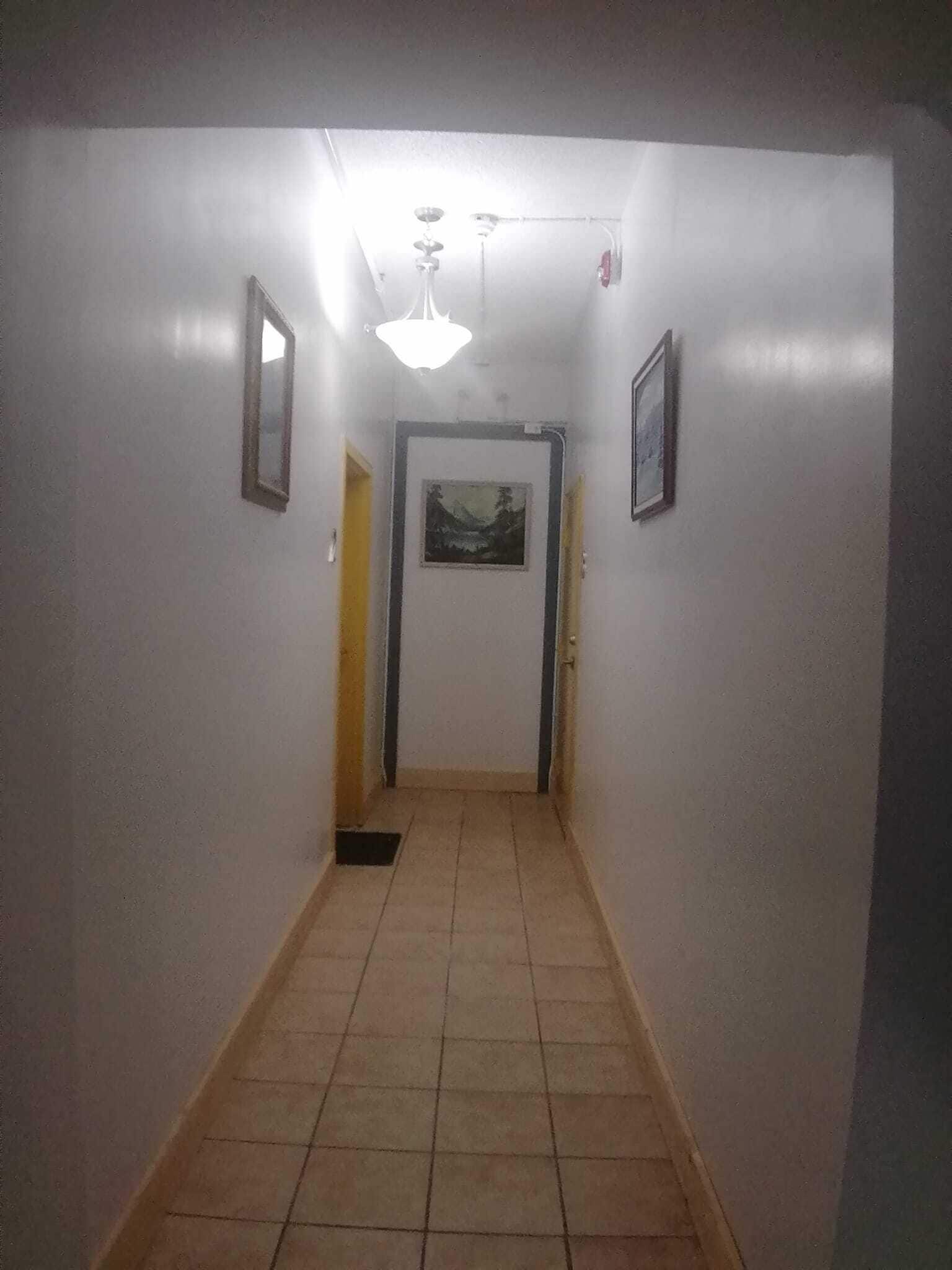 Un couloir étroit au sol carrelé, aux murs blancs et doté d’un ventilateur de plafond. Deux tableaux encadrés ornent les murs, et une porte se trouve au fond du couloir.