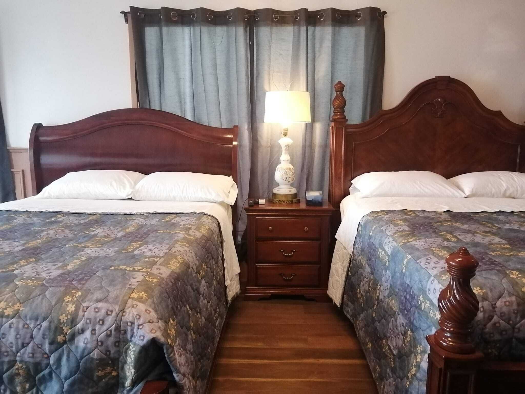 Una camera d'albergo con due letti in legno, un comodino con una lampada e una finestra con tende.