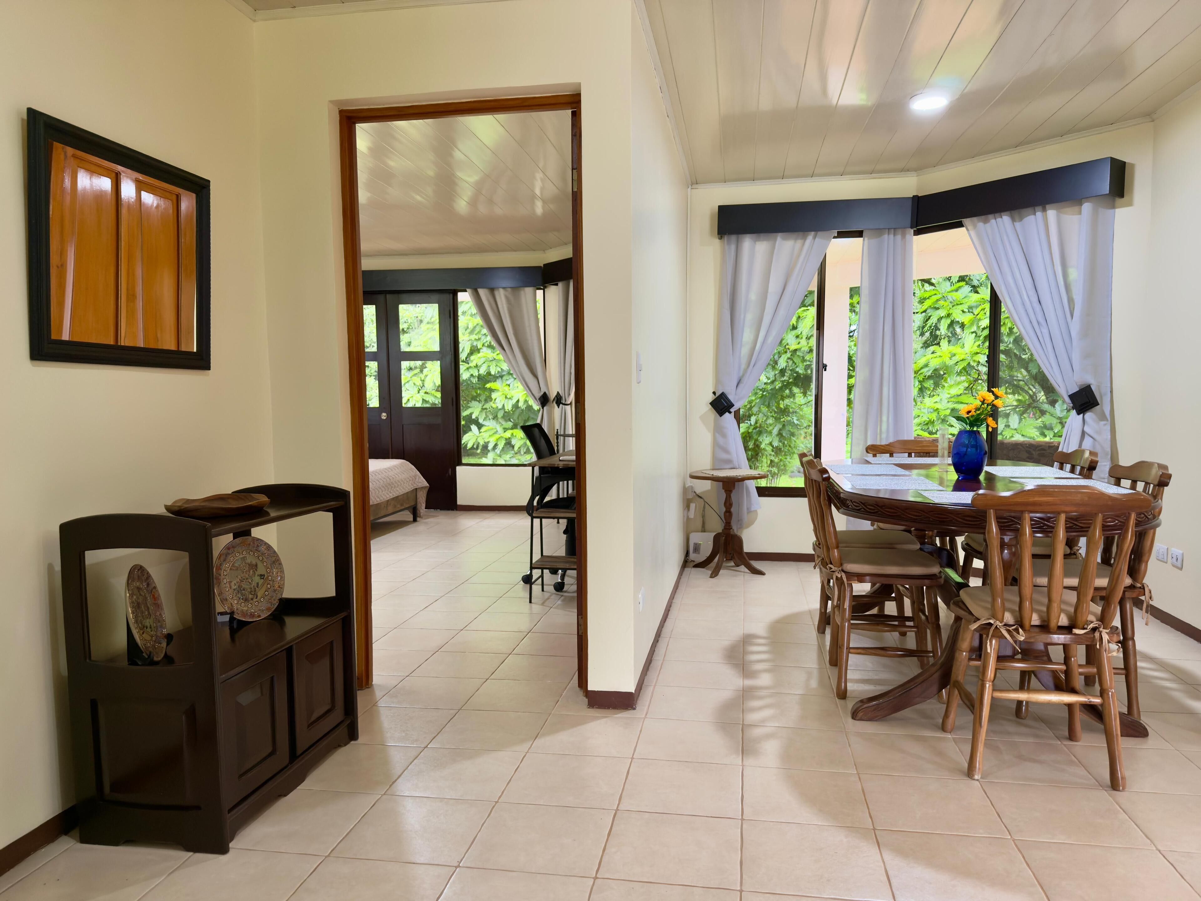Una habitación con una cama, una mesa de comedor con sillas, una mesita con una silla y un armario. Hay una puerta que da a otra habitación.
