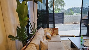 A modern living room with a long beige sofa, a coffee table, and a large window with a view of the outdoors. - A cozy  romantic private self contained room in the bushes of kitovu kitende  (Kampala)