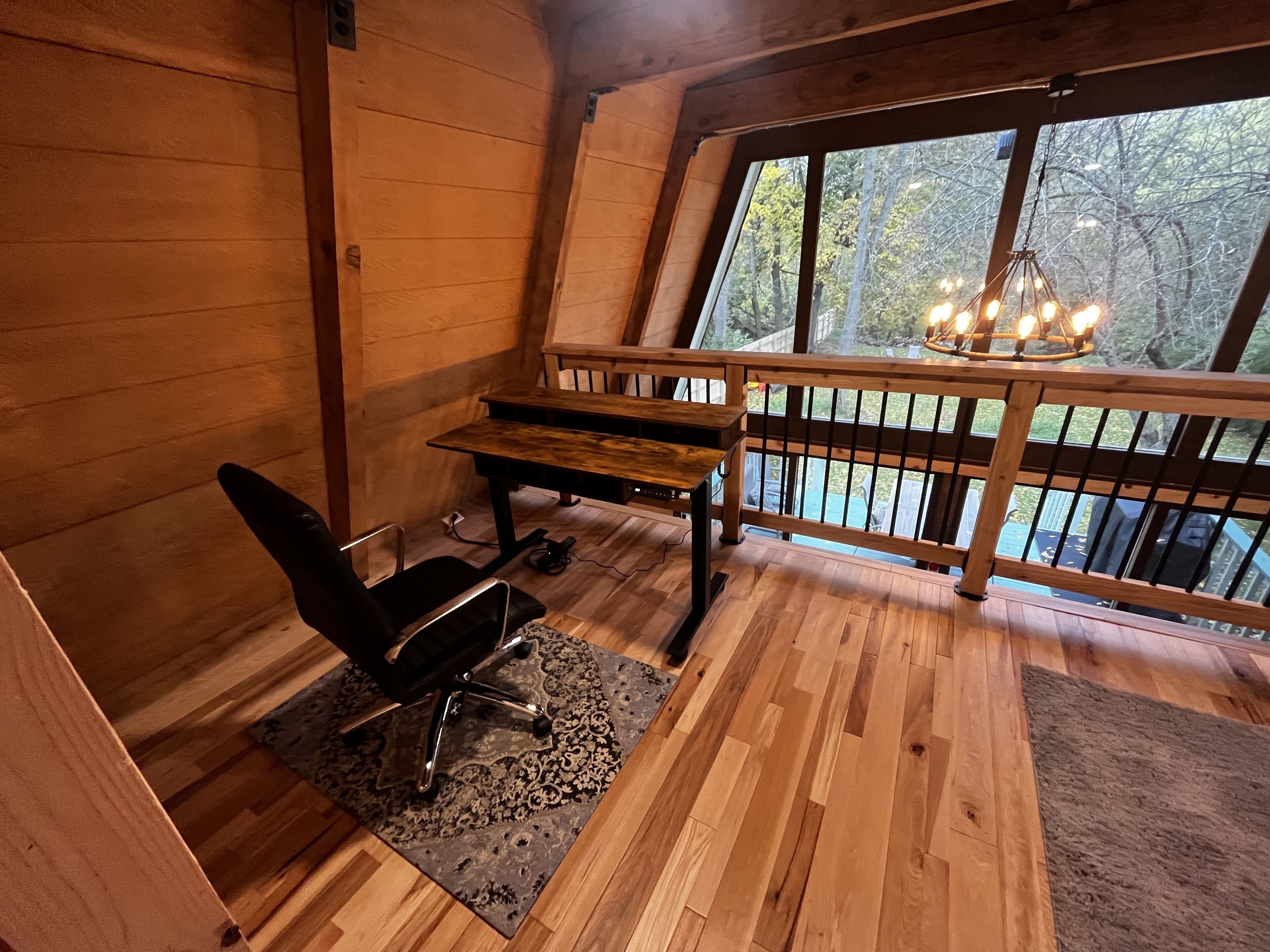 A cozy cabin room with a wooden desk, a chair, and a large window overlooking a forest.