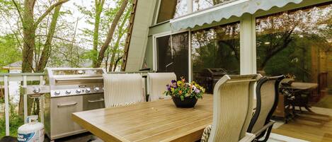 An outdoor patio with a wooden table, chairs, a grill, and a flower vase.