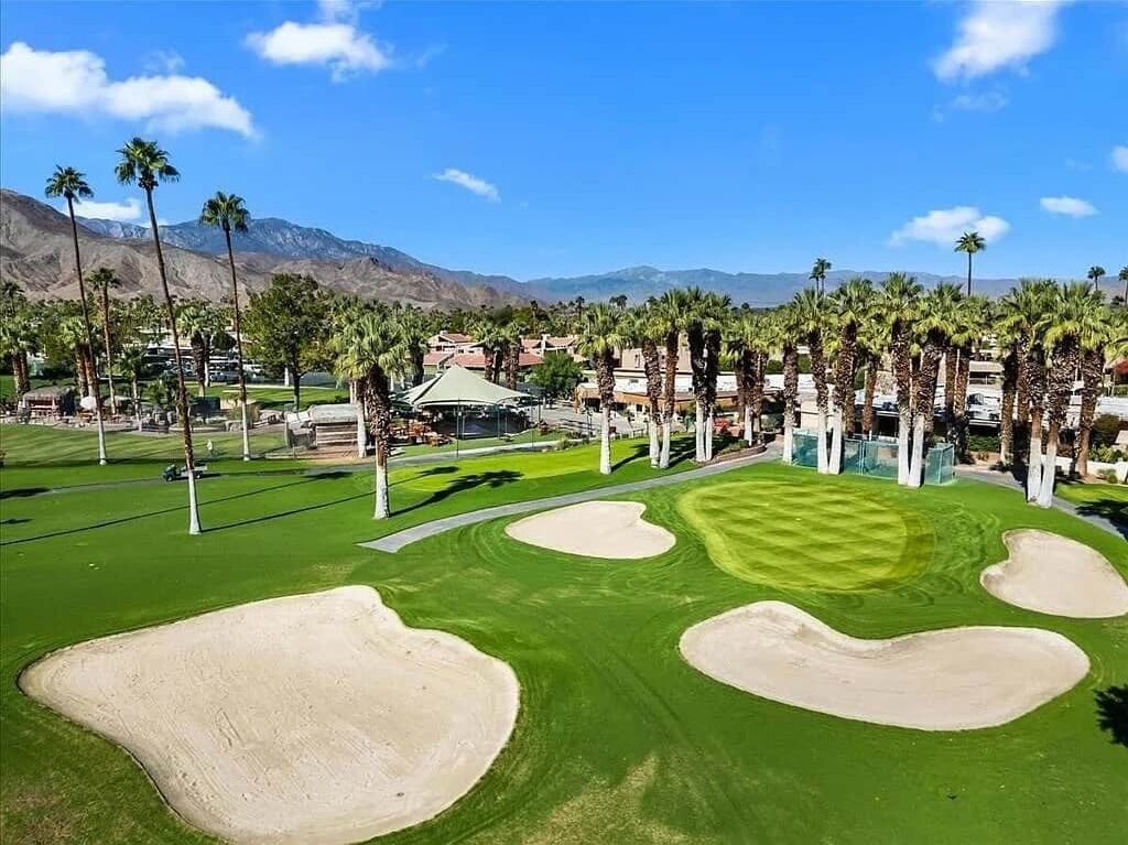 A golf course with sand bunkers, palm trees, and a mountainous backdrop.