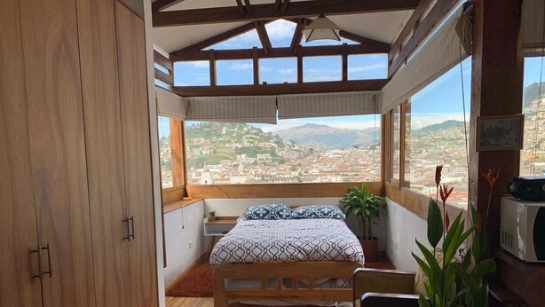 A bedroom with a wooden floor, a bed with patterned bedding, a small table, a potted plant, and a view of a cityscape through large windows. - Suite Centro Histórico (Quito)