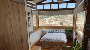 A bedroom with a wooden floor, a bed with patterned bedding, a window with a city view, and a potted plant. - Suite Centro Histórico (Quito)