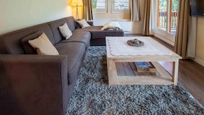 Una sala de estar con un sofá marrón, una mesa de centro de madera y vistas a una casa a través de ventanas amplias.