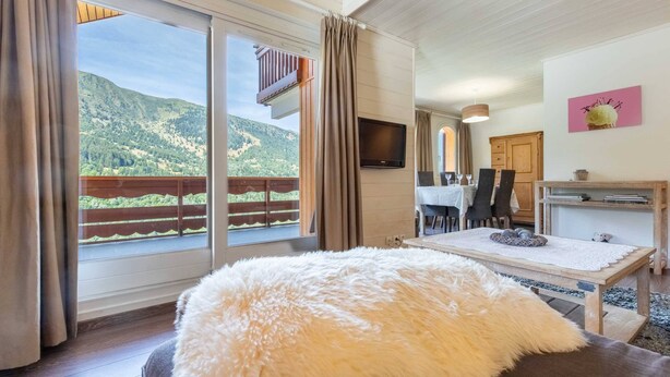 Una habitación con un ventanal grande, un otomano blanco y mullido, una mesa de madera y un televisor en la pared.