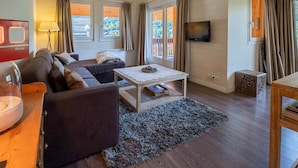 Una sala de estar acogedora con un sofá, una mesita de centro y un televisor. Se ven ventanas con cortinas, una lámpara y una pequeña mesa con un adorno.
