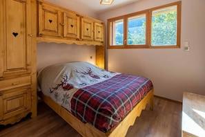 A wooden bed with a plaid blanket, a wooden wardrobe with heart-shaped cutouts, and a window with a view of greenery.