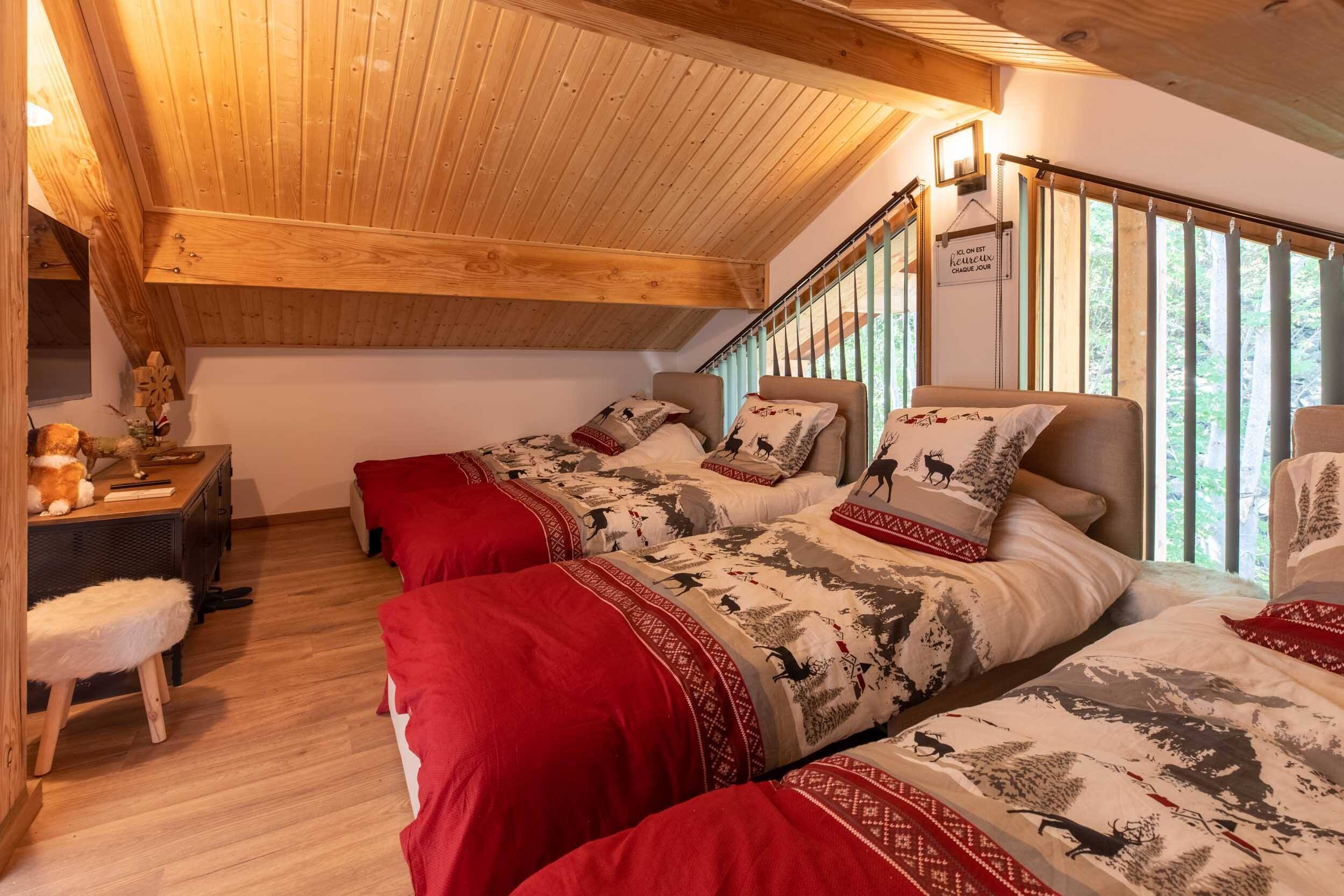 A cozy bedroom with three beds, wooden ceiling beams, a desk with a chair, and a window with curtains.