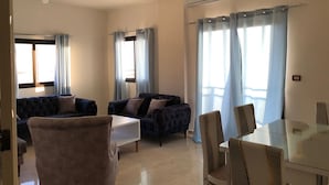 Ein Wohnzimmer mit einer Sofagarnitur, einem Esstisch und Stühlen. Es gibt ein Fenster mit Vorhängen, eine Klimaanlage an der Wand und eine Leuchte.