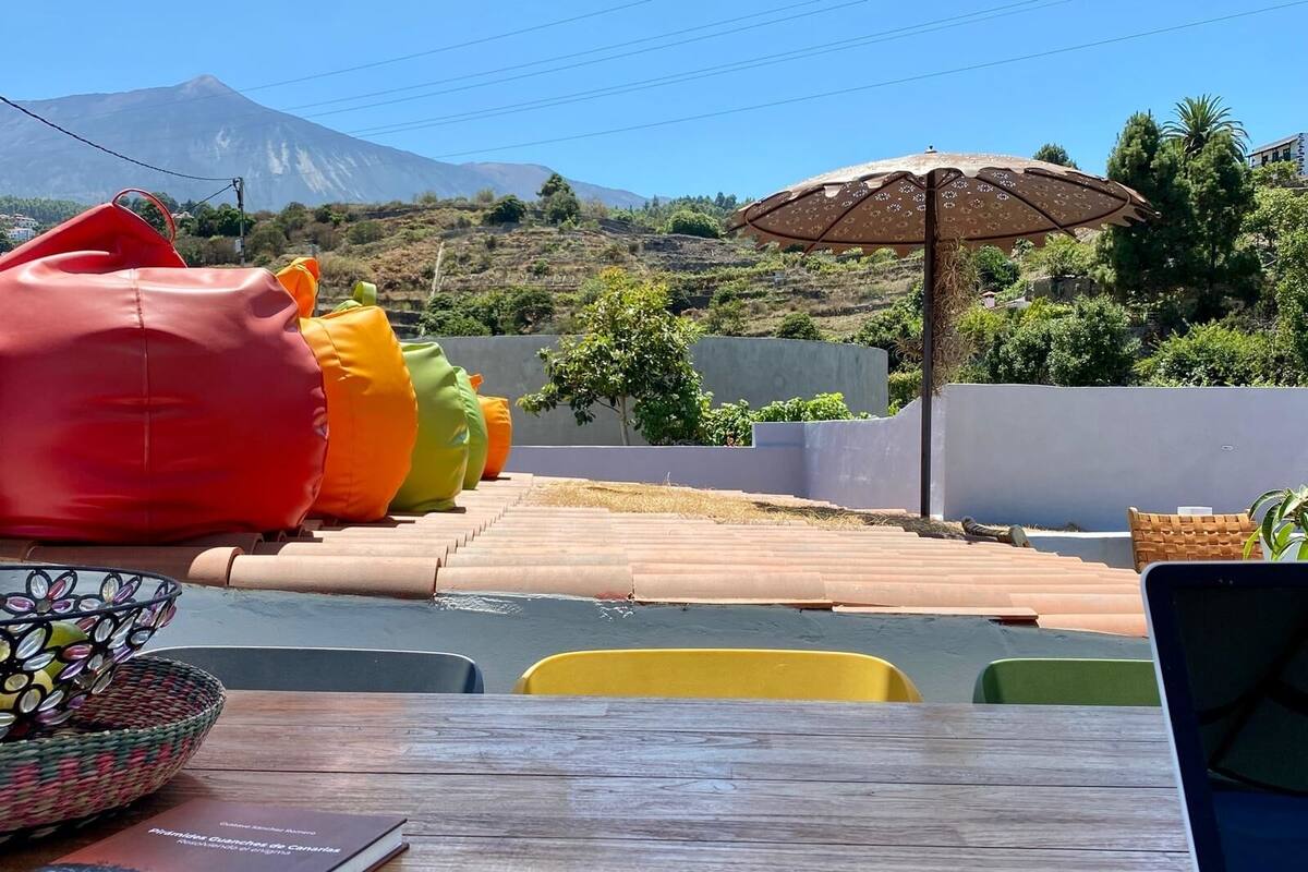 Una mesa con un libro, gafas de sol y un teléfono móvil. Se ven sillones de tía y un paraguas estampado al fondo.