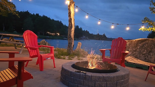 Ein Lagerfeuer mit roten Adirondack-Stühlen darum herum, mit Blick auf ein Gewässer.