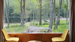 Ein Zimmer mit großem Fenster mit Blick auf einen Wald und einen See. Vor dem Fenster stehen ein Holztisch und zwei gelbe Stühle.