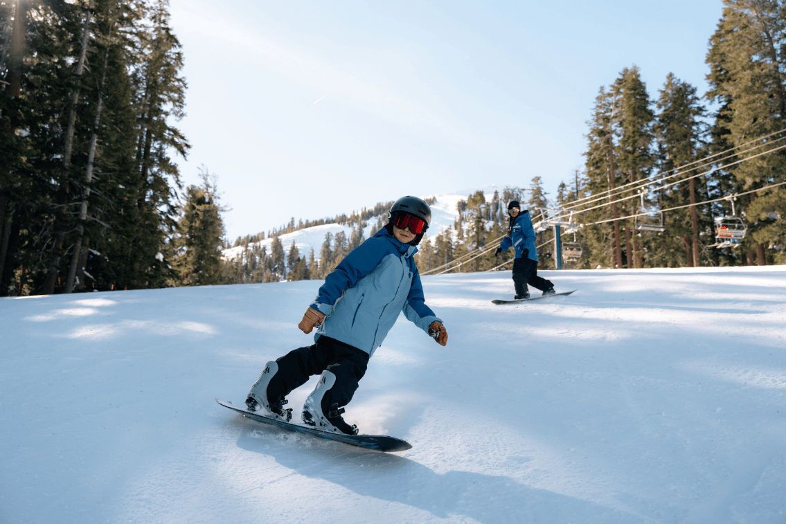 Two people snowboarding on a snowy slope with trees and ski lifts in the background.