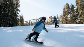 Two people snowboarding on a snowy slope with trees and ski lifts in the background.