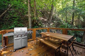 Une terrasse en bois avec un barbecue et une table de pique-nique, entourée d’une forêt dense.