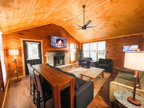 L’intérieur chaleureux d’un chalet avec un plafond en bois, une cheminée, un téléviseur fixé au-dessus, et un coin repas comprenant une table et des chaises.