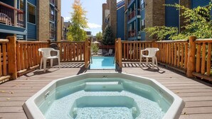 Un bain à remous sur une terrasse en bois, avec une piscine en arrière-plan, le tout entouré d’immeubles.