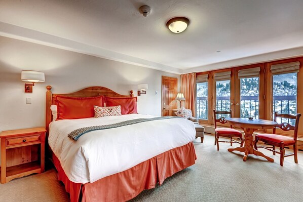 Habitación de hotel con una cama grande, cabecera de madera, mesa de comedor con sillas y vistas a un paisaje nevado a través de las ventanas.