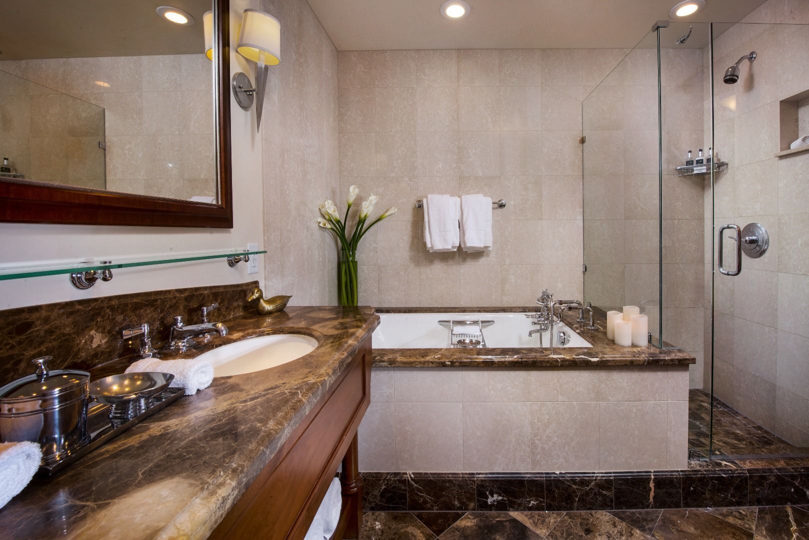 A bathroom with a marble vanity, a large bathtub, a glass-enclosed shower, and a mirror with wall sconces.
